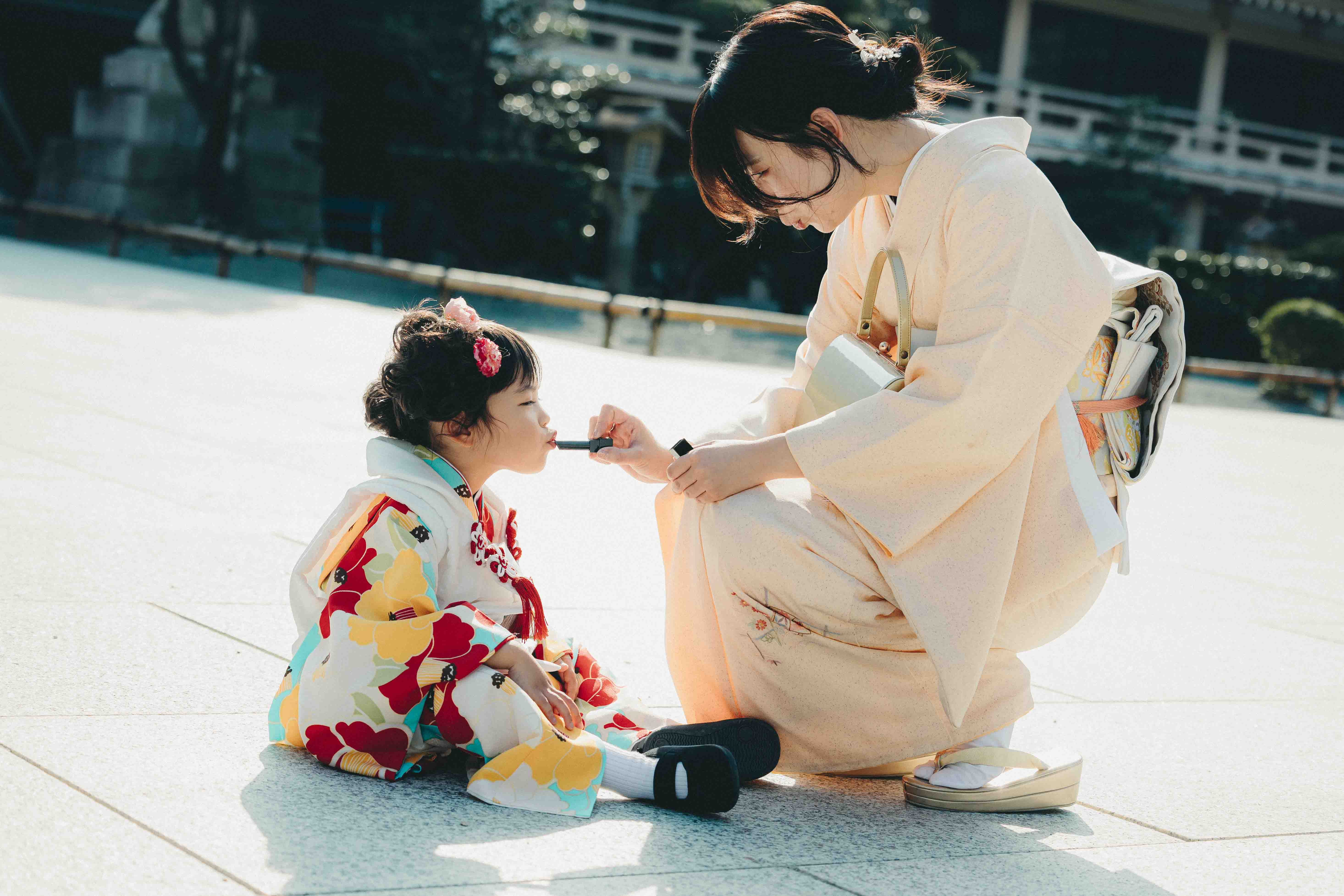七五三の着物姿の親子が寄り添い、母親が娘の唇にやさしく紅をさしている、やわらかな光に包まれた一場面