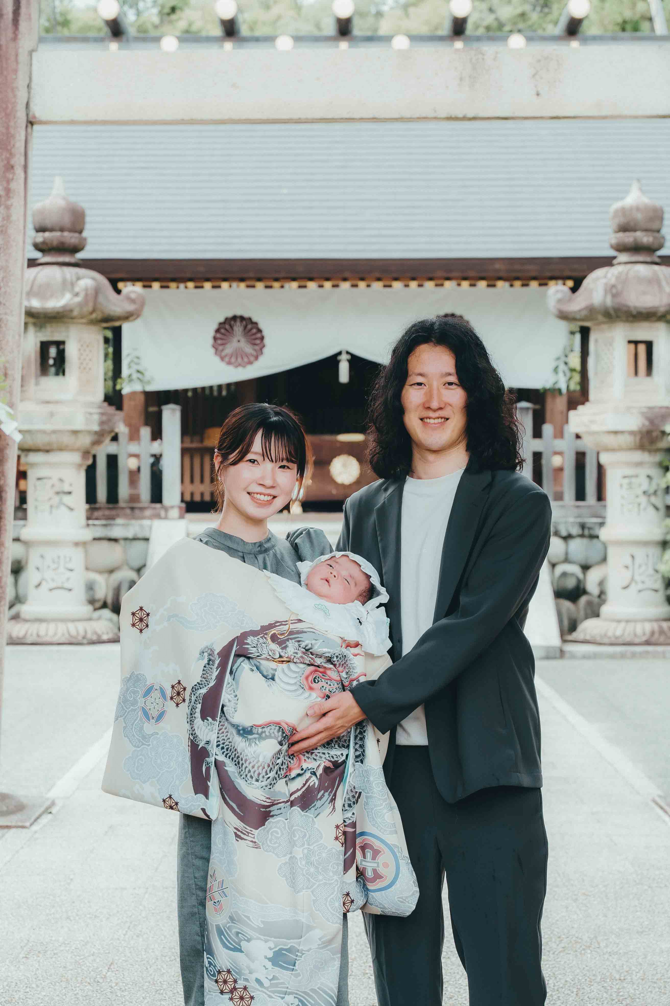 祝い着に包まれた赤ちゃんを抱く母親と、隣に寄り添う父親の記念写真
